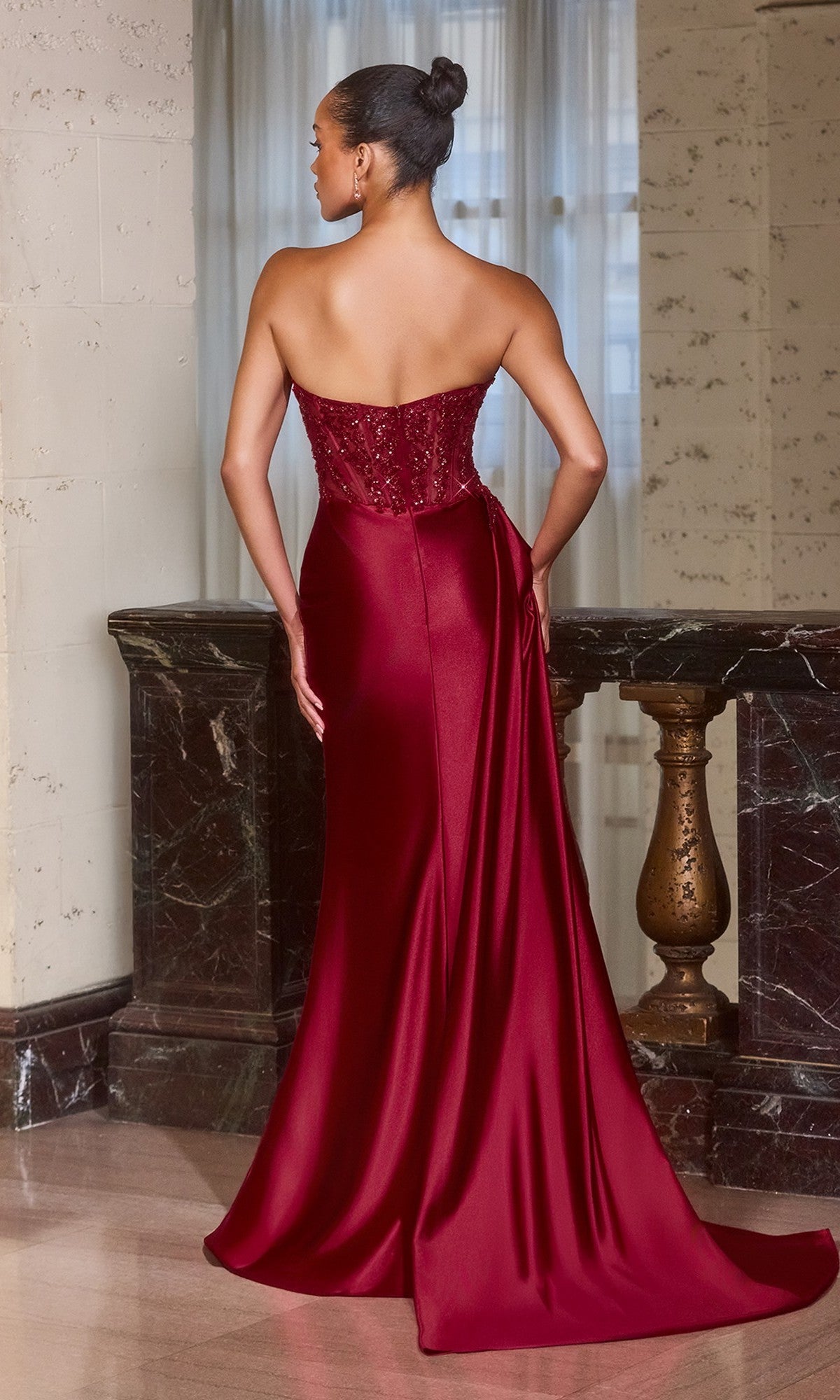 Woman wearing the Side-Drape Strapless Long Prom Gown (Ladivine J887), featuring a beaded bodice, satin floor-length skirt, and rich burgundy color, stands indoors with her back to the camera for an elegant formal look.